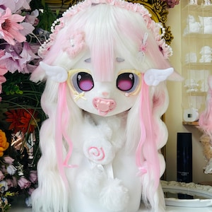 May include: A white and pink plush doll with large eyes, a pacifier, and a lollipop. The doll has white hair with pink streaks, angel wings, and a decorative headband. The doll is a whimsical, fantasy-themed item.