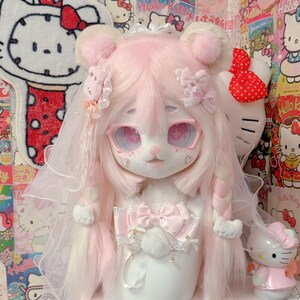 Könnte beinhalten: Eine Plüschpuppe mit rosa Haaren, großen Augen und einem Schleier. Die Puppe hat einen weißen Körper und ist mit Schleifen und Bändern verziert. Eine Hello Kitty-Figur und ein Hello Kitty-Teppich befinden sich im Hintergrund.