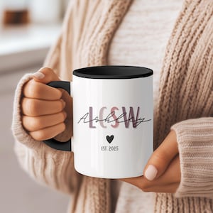 Peut inclure: Mug en céramique blanche avec intérieur et anse noirs. Le mug présente le nom "Ashley" en écriture cursive, avec les lettres "LCSW" superposées. Un cœur noir et le texte "EST. 2025" sont également présents.