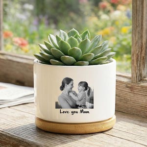 Puede incluir: Una maceta de cerámica blanca con una planta suculenta. La maceta presenta una foto en blanco y negro de una madre y un niño, con el texto "Love you Mam" debajo. Se encuentra sobre una base redonda de madera.