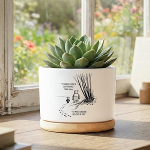 May include: A white ceramic plant pot with a succulent. The pot features a black and white illustration of Winnie the Pooh and Piglet with the quote "IT MAKES SUCH A DIFFERENCE SAID Asher TO MAKE SOMEONE BELIEVE IN YOU". The pot sits on a wooden base.