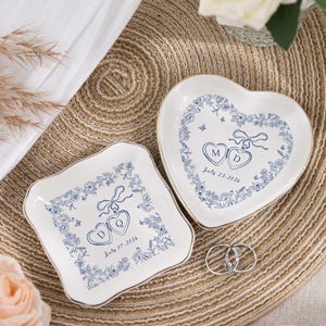May include: Two white ceramic ring dishes, one heart-shaped and one square, with blue floral designs and gold trim. Each dish has initials and a date. Two silver rings are placed on the woven mat.