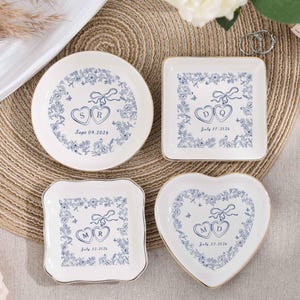 May include: Four white ceramic ring dishes with blue floral designs and gold rims. Each dish features initials and a date. Shapes include round, square, and heart. The text on the dishes reads "SR Sept 09, 2026", "DQ July 27, 2026", "MR July 22, 2026", and "MD July 27, 2026".