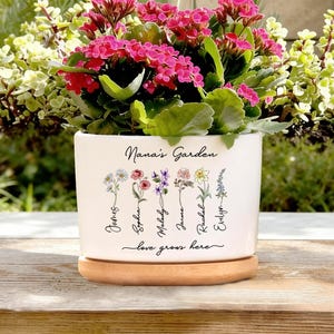 May include: A white ceramic plant pot with a wooden base, filled with vibrant pink flowers. The pot is inscribed with "Nana's Garden" and features floral illustrations with names. The phrase "love grows here" is also written on the pot.