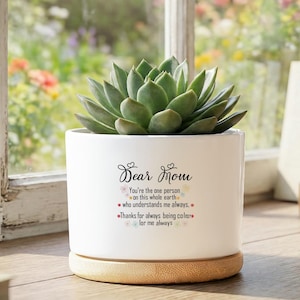 Puede incluir: Una maceta de cerámica blanca con una planta suculenta. La maceta tiene el texto "Dear Mom" y un mensaje. La maceta se encuentra sobre una base de madera, cerca de una ventana.