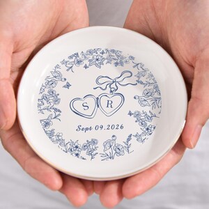May include: A white ceramic ring dish with a gold rim, held in two hands. The dish features a blue floral design around the edge and two heart-shaped lockets with the initials "S" and "R". The date "Sept 09.2026" is printed below.