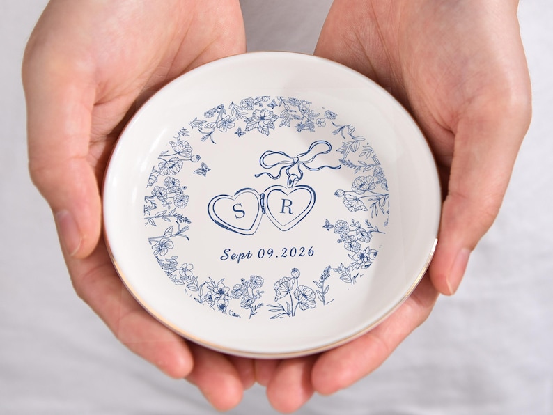 May include: A white ceramic ring dish with a gold rim, held in two hands. The dish features a blue floral design around the edge and two heart-shaped lockets with the initials "S" and "R". The date "Sept 09.2026" is printed below.