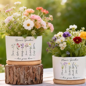 May include: Two white ceramic flower pots with colorful flowers. One pot reads "Nana's Garden" and the other reads "Mom's Garden". Each pot has names and the phrase "love grows here" written on it. The pots sit on wooden bases.