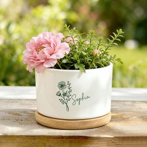 May include: A white ceramic plant pot with a light brown wooden coaster, containing a pink flower and green leaves. The pot features a floral design and the name "Sophia" in green script. The background is a blurred green.