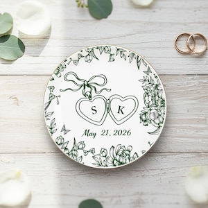 Może przedstawiać: Biały ceramiczny talerz ze złotą obwódką, z zielonym kwiatowym wzorem i dwoma medalionami w kształcie serca z inicjałami "S" i "K". Wyświetlana jest również data "21 maja 2026". Widoczne są dwa złote obrączki.