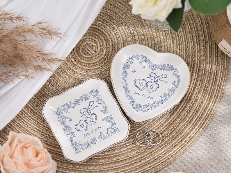 May include: Two white ceramic ring dishes, one heart-shaped and one square, with blue floral designs and gold trim. Each dish has initials and a date. Two silver rings are placed on the woven mat.