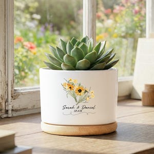 Könnte beinhalten: Ein weißer Keramik-Pflanztopf mit einer Sukkulentenpflanze. Der Topf hat ein Blumendesign mit gelben Sonnenblumen und den Namen "Sarah & Daniel" mit dem Datum "10.8.26" in Schrift. Er steht auf einem runden, hellen Holzsockel.