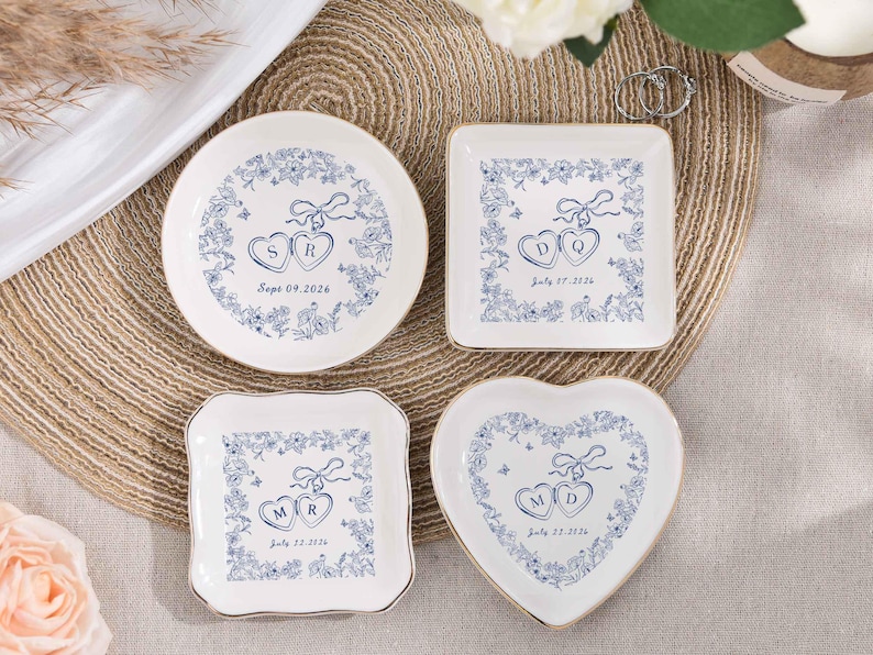 May include: Four white ceramic ring dishes with blue floral designs and gold rims. Each dish features initials and a date. Shapes include round, square, and heart. The text on the dishes reads "SR Sept 09, 2026", "DQ July 27, 2026", "MR July 22, 2026", and "MD July 27, 2026".