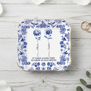 Könnte beinhalten: Ein quadratisches, weißes Keramik-Schmucktablett mit blauem Blumendesign und goldenem Rand. Das Tablett zeigt zwei stilisierte Blumen mit den Namen "Olivia" und "Sophia" und dem Text "IT TAKES A LONG TIME TO GROW AN OLD FRIEND."
