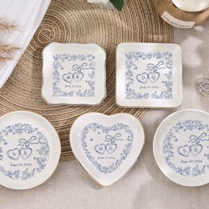 May include: A selection of white ceramic ring dishes in various shapes: square, round, and heart-shaped. Each dish features a blue floral design with initials and a date. The dishes have gold rims and are arranged on a neutral surface.