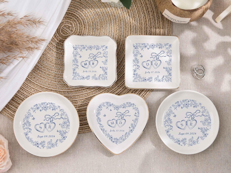 May include: A selection of white ceramic ring dishes in various shapes: square, round, and heart-shaped. Each dish features a blue floral design with initials and a date. The dishes have gold rims and are arranged on a neutral surface.