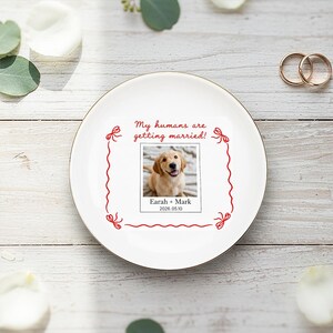 May include: A white ceramic plate with a gold rim, displaying a photograph of a golden retriever puppy. The plate has red text saying "My humans are getting married!" and the names "Earah + Mark" with the date "2026.05.10". Two gold wedding rings are present.