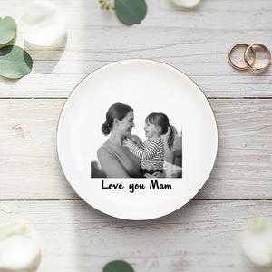 Op de afbeelding: Een witte keramische plaat met een gouden rand toont een zwart-witfoto van een moeder en kind. De tekst "Love you Mam" is eronder gedrukt. Twee gouden ringen liggen in de buurt van de plaat.