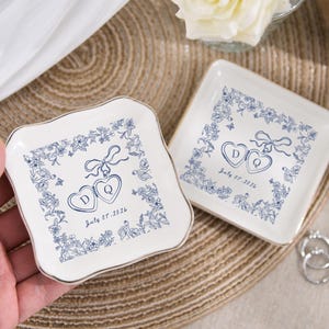 May include: Two white ceramic ring dishes with blue floral and heart designs. Each dish has a gold rim and features the initials "D" and "Q" inside hearts, with the date "July 07, 2026".