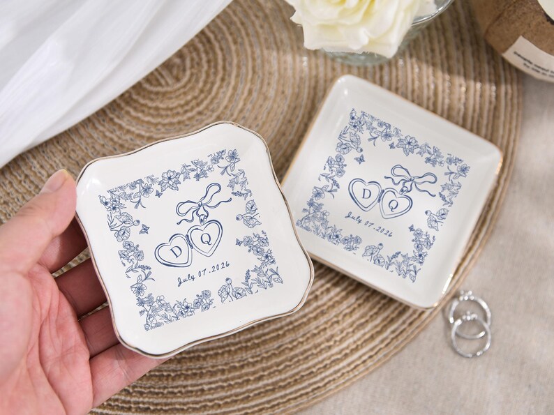May include: Two white ceramic ring dishes with blue floral and heart designs. Each dish has a gold rim and features the initials "D" and "Q" inside hearts, with the date "July 07, 2026".