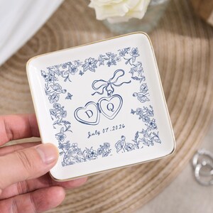 May include: Square white ceramic ring dish with a gold rim. The dish features a blue floral design around the edges and two heart-shaped lockets with the letters "D" and "Q". The date "July 07.2026" is written below the lockets.