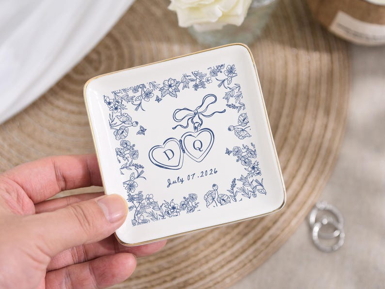May include: Square white ceramic ring dish with a gold rim. The dish features a blue floral design around the edges and two heart-shaped lockets with the letters "D" and "Q". The date "July 07.2026" is written below the lockets.