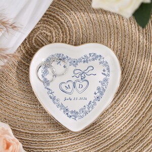 May include: A heart-shaped white ceramic ring dish with a blue floral design and gold trim. The dish features the initials "M" and "D" inside hearts, with the date "July 21, 2026". Two silver hoop earrings rest on the dish.