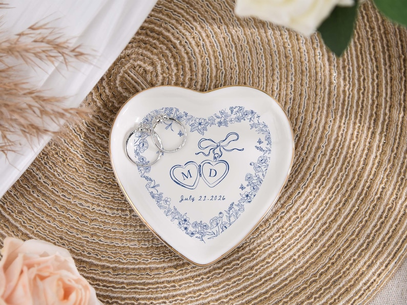 May include: A heart-shaped white ceramic ring dish with a blue floral design and gold trim. The dish features the initials "M" and "D" inside hearts, with the date "July 21, 2026". Two silver hoop earrings rest on the dish.
