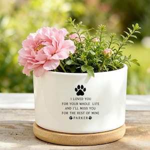 Puede incluir: Una maceta de cerámica blanca con una flor rosa y follaje verde. La maceta tiene una huella de pata y el texto "I LOVED YOU FOR YOUR WHOLE LIFE AND I'LL MISS YOU FOR THE REST OF MINE PARKER". La maceta está sobre un posavasos de madera.