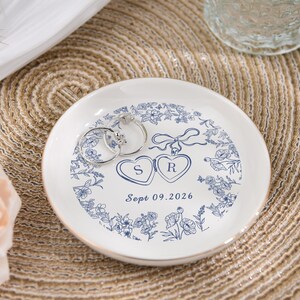 May include: A white ceramic ring dish with a blue floral pattern and gold edging. Two silver rings are placed on the dish. The dish has two hearts with the letters "S" and "R" and the date "Sept 09.2026".