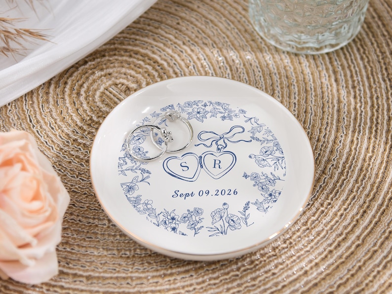 May include: A white ceramic ring dish with a blue floral pattern and gold edging. Two silver rings are placed on the dish. The dish has two hearts with the letters "S" and "R" and the date "Sept 09.2026".