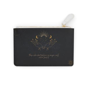 Pode incluir: Bolsa de mão preta com zíper dourado e puxador branco. Apresenta uma ilustração dourada de mãos, luas e um sol, com o texto "Aqueles que não acreditam em magia, nunca a encontrarão."