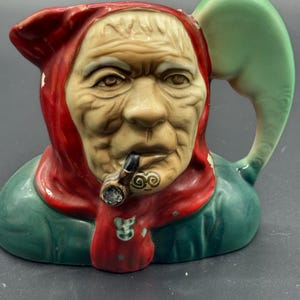 May include: A ceramic pitcher with a detailed face of an elderly person wearing a red hood and scarf. The pitcher has a green handle and base, and the person is smoking a pipe.
