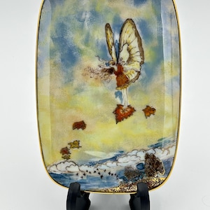 Könnte beinhalten: Rechteckige dekorative Platte mit goldenem Rand. Die Platte zeigt eine skurrile Illustration einer Fee mit Schmetterlingsflügeln, die über einer Landschaft mit fallenden Blättern, Bäumen und Wolken schwebt. Der Hintergrund hat einen Farbverlauf von Gelb und Blau.
