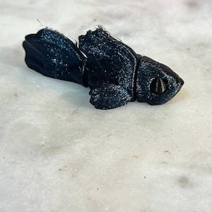 Peut inclure: Petite figurine de poisson bleu foncé imprimée en 3D. Le poisson présente un design détaillé avec une texture visible et un petit œil rond. La figurine est posée sur une surface claire, peut-être en marbre.