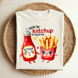 May include: Off-white t-shirt featuring a cartoon ketchup bottle and a box of fries, both with smiling faces. The text "You're the ketchup to my fries" is printed above the illustrations. The shirt is on a woven basket.
