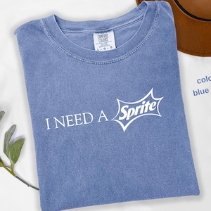 May include: Blue jean colored t-shirt with the text "I NEED A" and the Sprite logo in white. The shirt is folded and has a crew neck. A brown hat is partially visible in the background.