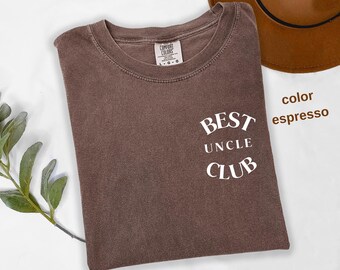 Puede incluir: Camiseta color espresso con el texto blanco "BEST UNCLE CLUB". La camiseta es de la marca Comfort Colors. Un sombrero marrón es visible en el fondo. La camiseta está doblada sobre una superficie blanca.