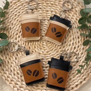 Puede incluir: Cuatro llaveros con forma de taza de caf&eacute;. Cada llavero est&aacute; hecho de cuero en tonos beige, marr&oacute;n y negro. La secci&oacute;n marr&oacute;n presenta dos dise&ntilde;os de granos de caf&eacute;. Los llaveros est&aacute;n colocados sobre un salvamanteles redondo y tejido.