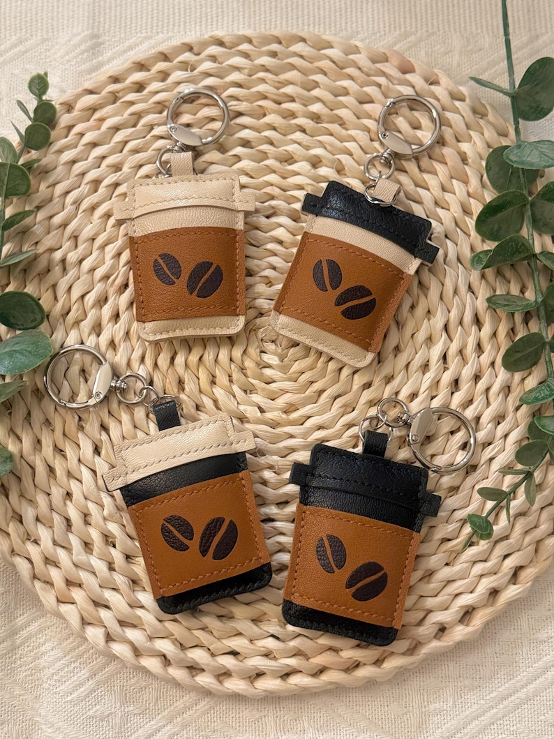 Puede incluir: Cuatro llaveros con forma de taza de caf&eacute;. Cada llavero est&aacute; hecho de cuero en tonos beige, marr&oacute;n y negro. La secci&oacute;n marr&oacute;n presenta dos dise&ntilde;os de granos de caf&eacute;. Los llaveros est&aacute;n colocados sobre un salvamanteles redondo y tejido.
