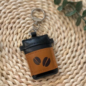 Puede incluir: Un llavero de cuero negro y marr&oacute;n con forma de taza de caf&eacute;. El llavero tiene un anillo plateado y una banda marr&oacute;n con dos dise&ntilde;os de granos de caf&eacute;. El llavero est&aacute; sobre una superficie tejida.