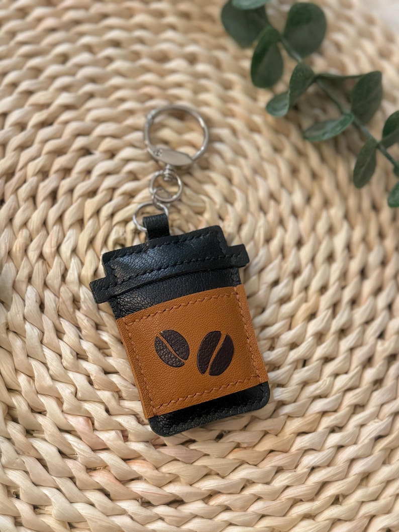 Puede incluir: Un llavero de cuero negro y marr&oacute;n con forma de taza de caf&eacute;. El llavero tiene un anillo plateado y una banda marr&oacute;n con dos dise&ntilde;os de granos de caf&eacute;. El llavero est&aacute; sobre una superficie tejida.
