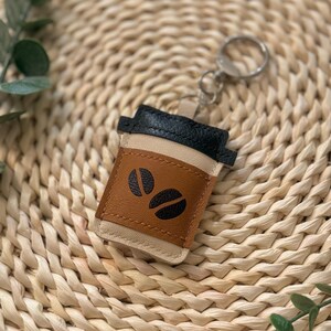Puede incluir: Un llavero de taza de caf&eacute; de cuero marr&oacute;n, negro y beige. El llavero presenta un dise&ntilde;o de taza de caf&eacute; con dos gr&aacute;ficos de granos de caf&eacute;. Incluye un anillo de metal plateado para sujetarlo a llaves o a un bolso.