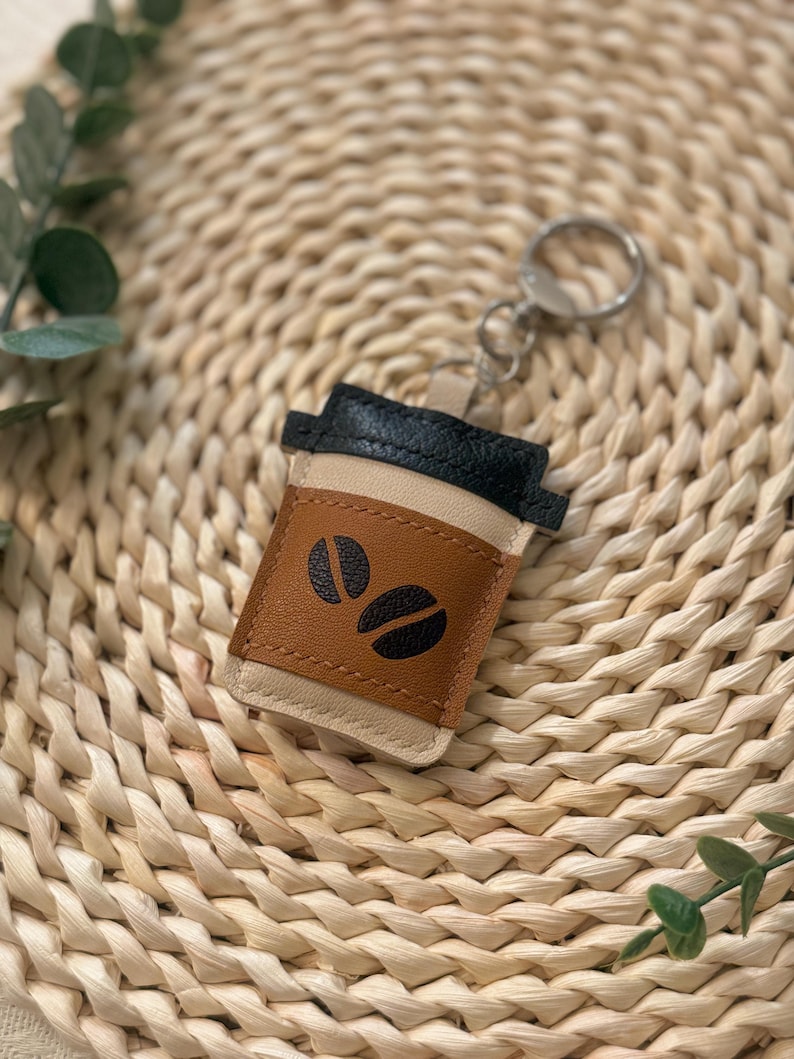 Puede incluir: Un llavero de taza de caf&eacute; de cuero marr&oacute;n, negro y beige. El llavero presenta un dise&ntilde;o de taza de caf&eacute; con dos gr&aacute;ficos de granos de caf&eacute;. Incluye un anillo de metal plateado para sujetarlo a llaves o a un bolso.