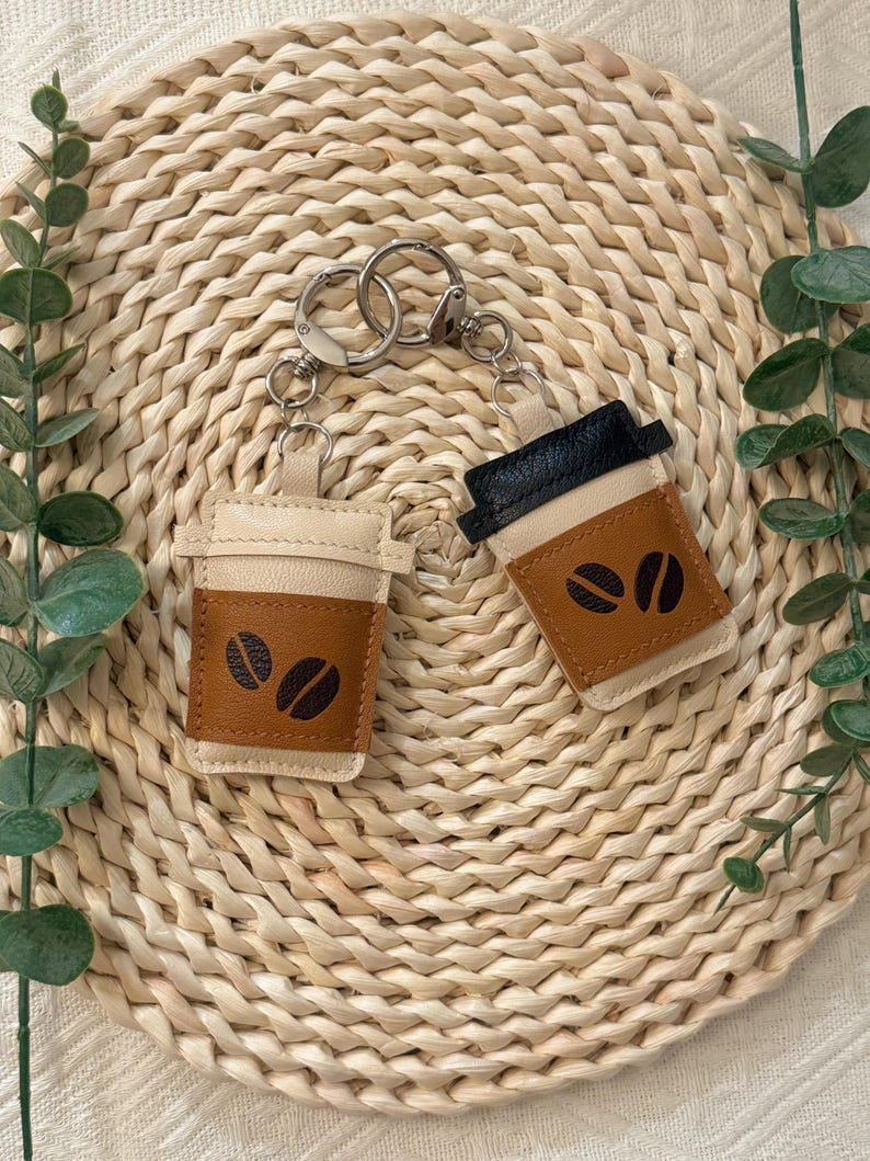 Puede incluir: Dos llaveros con forma de taza de caf&eacute;. Un llavero es de color beige y marr&oacute;n con detalles de granos de caf&eacute; negros. El otro llavero es negro y marr&oacute;n con detalles de granos de caf&eacute; negros. Ambos tienen llaveros plateados.