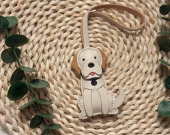 Puede incluir: Un llavero de cuero color crema con forma de perro, con orejas marrones y un lazo de cuero marrón. El perro tiene ojos negros, nariz negra y lengua roja. El llavero está sobre un salvamanteles circular tejido con hojas verdes.