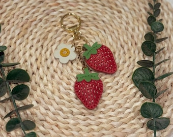 Puede incluir: Un llavero dorado con dos dijes de fresas rojas y un dije de flor blanca. Las fresas tienen copas verdes y están detalladas con pequeños patrones cosidos. El llavero está sobre un fondo circular tejido.