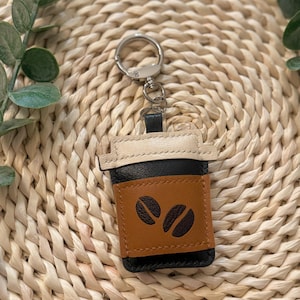 Puede incluir: Un llavero de cuero con forma de taza de caf&eacute;. El llavero presenta una funda marr&oacute;n con dos dise&ntilde;os de granos de caf&eacute;, una taza negra y una tapa color crema. Est&aacute; unido a un llavero plateado.