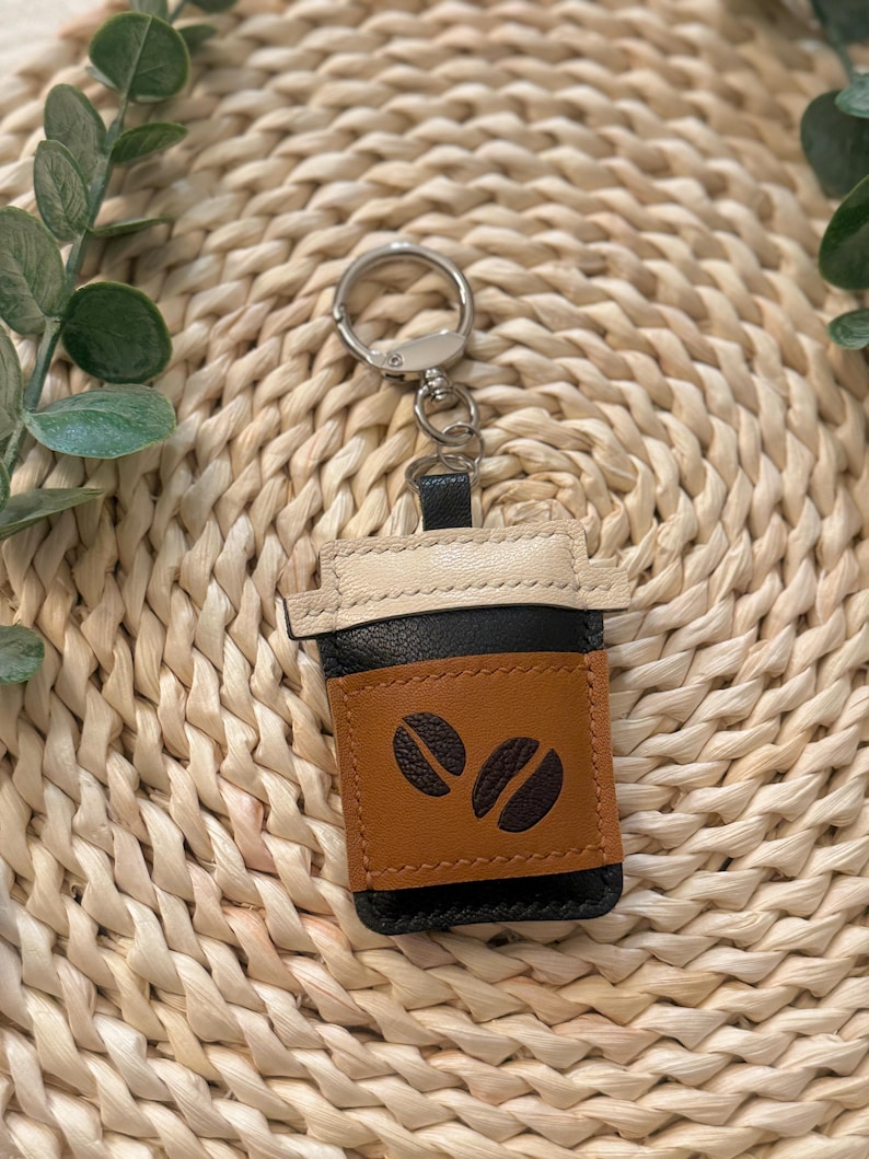 Puede incluir: Un llavero de cuero con forma de taza de caf&eacute;. El llavero presenta una funda marr&oacute;n con dos dise&ntilde;os de granos de caf&eacute;, una taza negra y una tapa color crema. Est&aacute; unido a un llavero plateado.