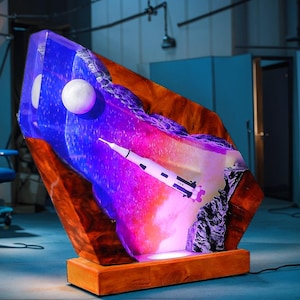 May include: A unique, illuminated sculpture featuring a space-themed design. The artwork showcases a rocket, a moon, and a vibrant display of purple and blue hues. The sculpture is mounted on a wooden base.